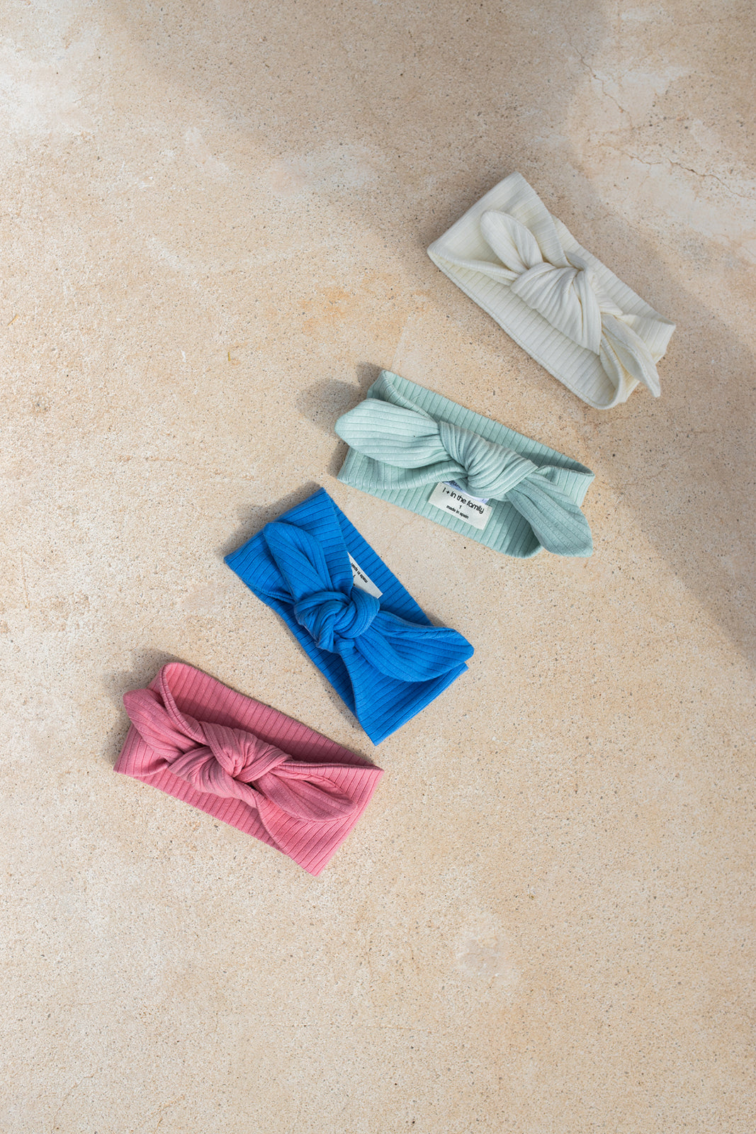 1+ in the family Ametlla off-white drapiert auf beigefarbenem Steinboden mit drei weiteren Stirnbändern in grün, blau und rot