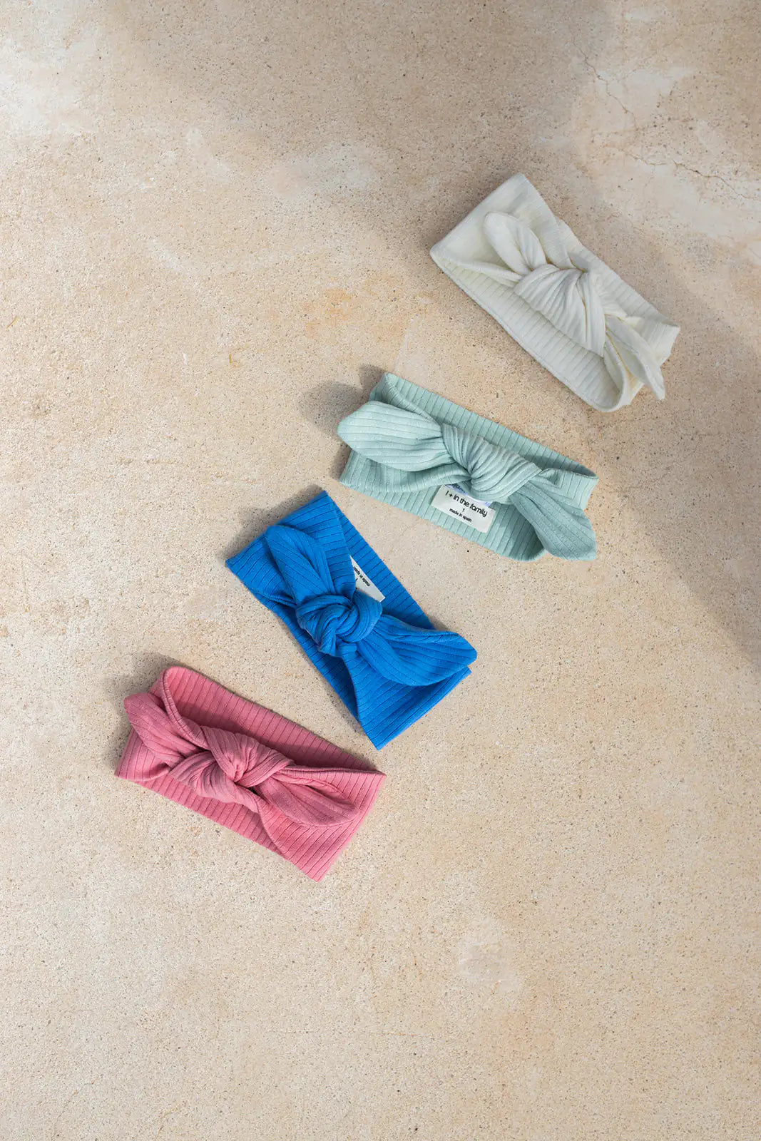 1+ in the family Ametlla off-white drapiert auf beigefarbenem Steinboden mit drei weiteren Stirnbändern in grün, blau und rot