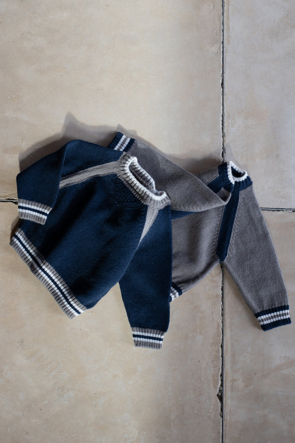 1 in the family Pullover Milano blue drapiert mit dem Pullover Milano in dark taupe auf einem beigefarbenen Fliesenboden. 