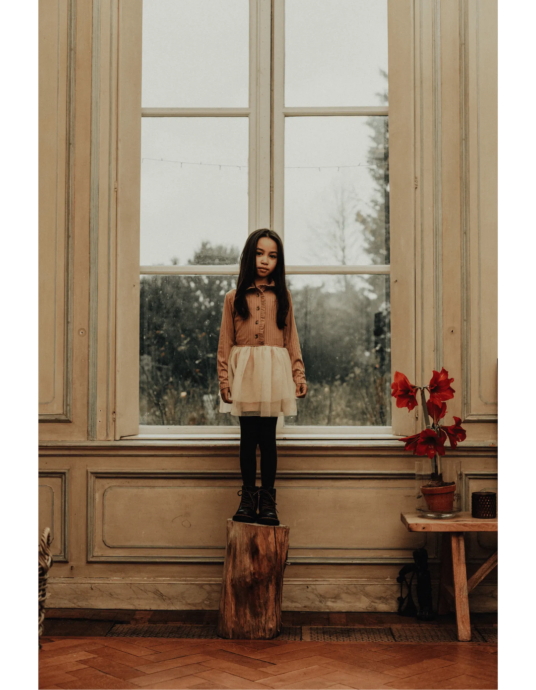 Baje Kleid Marly peach almond getragen von einem Mädchen stehend vor einem Fenster. 