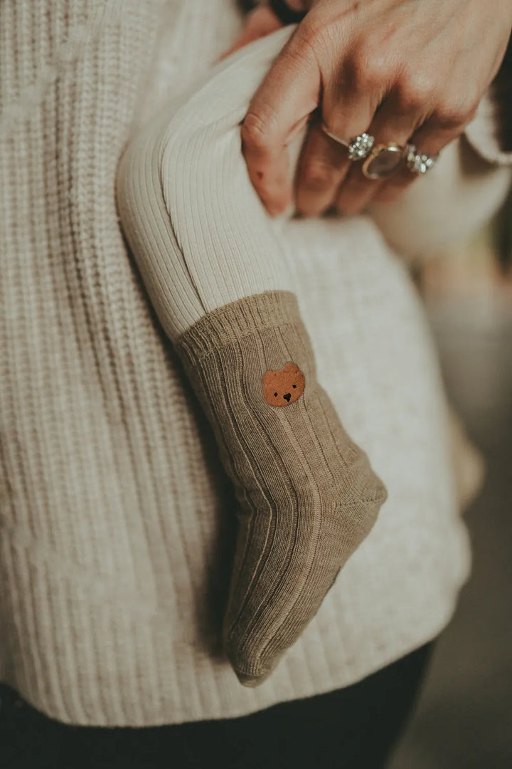 Donsje Bell Socks Bear dusty green getragen von einem Baby, das sich auf dem Arm einer erwachsenen Person befindet mit Fokus auf der Socke mit dem Bärengesicht