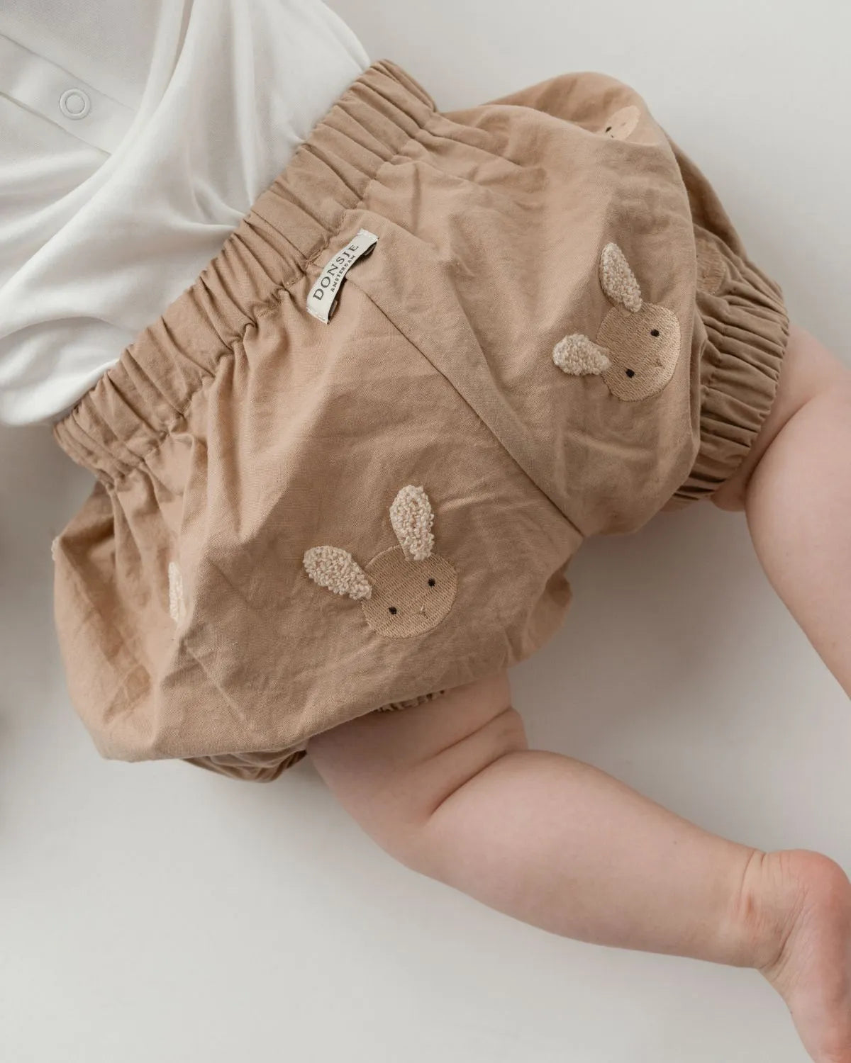Donsje Carson Bloomers Fluffy Bunnies rose mocha getragen von einem auf dem Bauch liegenden Baby mit Sicht auf die hinten aufgestickten Hasen und das angenähte Label von Donsje