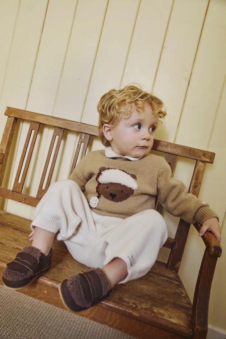 Donsje Denn Merino Wool Sweater Christmas Bear latte melange getragen von einem Jungen auf einer Holzbank sitzend, Ansicht von vorne. 