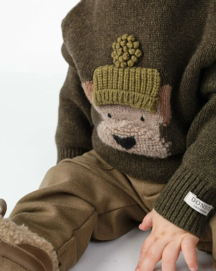 Donsje Denn Merino Wool Sweater Terrier dark olive getragen von einem sitzenden Baby, Ansicht von vorne. 