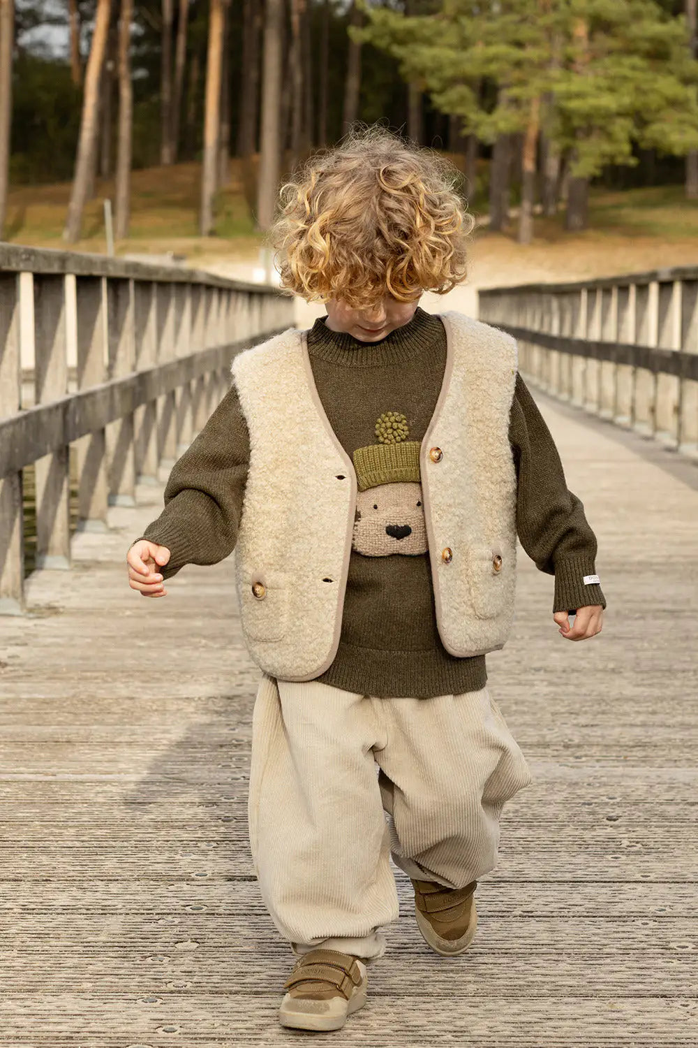 Donsje Denn Merino Wool Sweater Terrier dark olive getragen von einem Kind, das über eine Brücke läuft. Ansicht von vorne. 