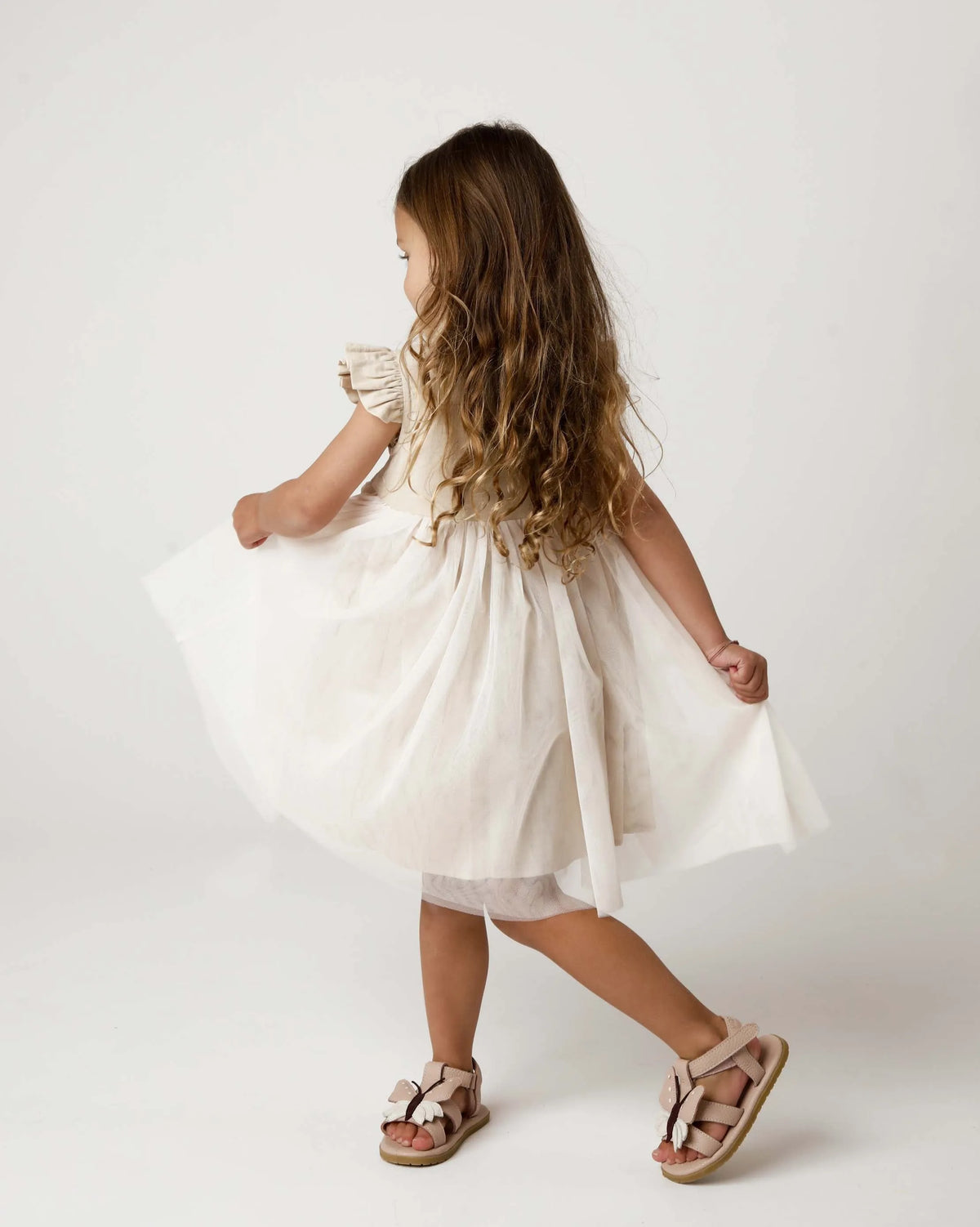 Donsje_Fleurance_Dress_soft_sand getragen von einem Mädchen vor weißem Hintergrund, das sich gerade zur rechten Seite dreht und dabei den Tüllrock schwingt