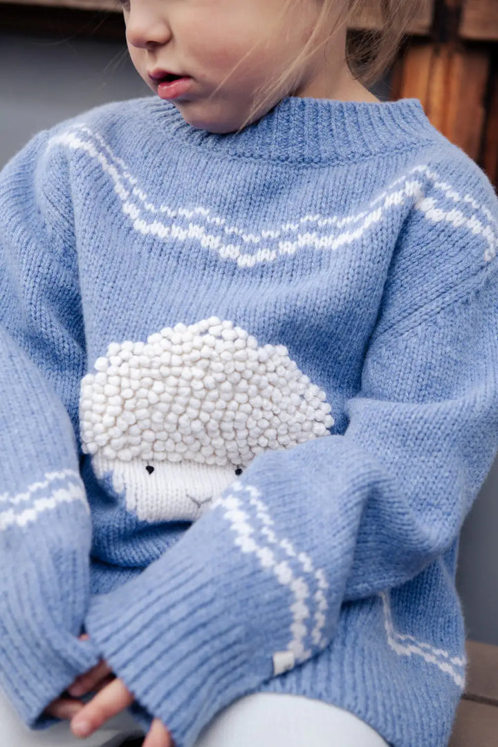 Donsje Selwyn Merino Wool Sweater Lammy vintage blue melange getragen von einem kleinen Mädchen, Detailansicht eingestricktes Lamm-Gesicht, Ansicht von vorne. 