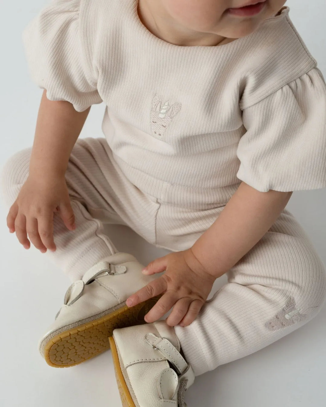 Donsje Soenie Leggings Unicorn soft powder und Donsje Nowien Top unicorn soft powder getragen von einem sitzenden Baby mit Blick auf die Leggings mit dem Einhorn-Gesicht und dem gleichfarbigen T-Shirt