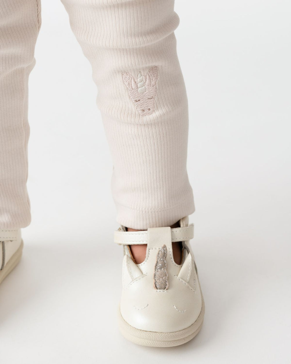 Donsje Soenie Leggings Unicorn soft powder getragen von einem stehenden Kleinkind vor weißem Hintergrund mit Detailaufnahme vom Einhorn-Gesicht 