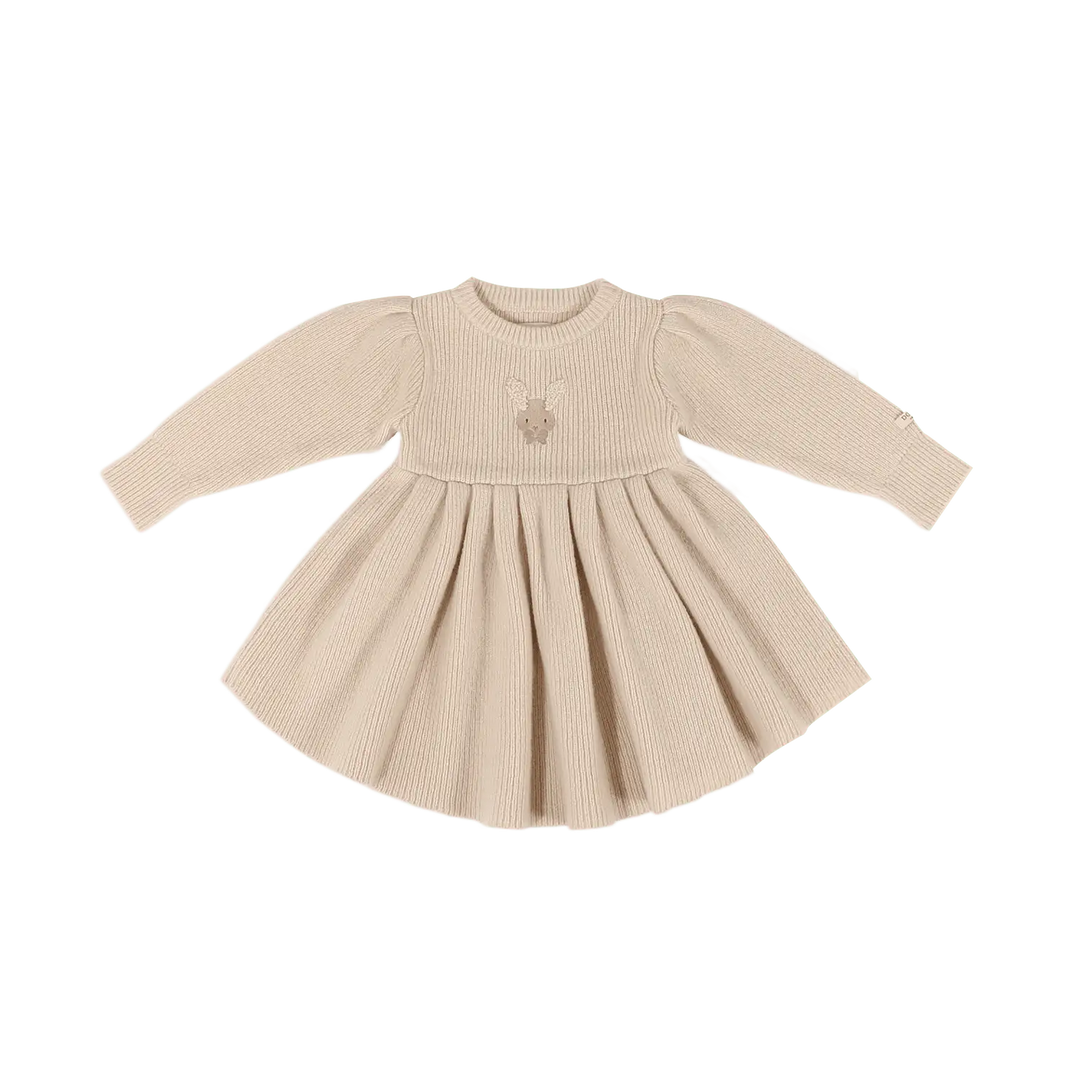 cremefarbenes, langärmeliges Kleid für Baby-Mädchen aus 100% Merinowolle in feiner, gerippter Struktur mit einem aufgestickten Hasengesicht mit flauschigen Ohren und einer rosafarbenen Schleife auf Höhe der Brust, Rundhalsausschnitt, A-Linie, leichte Raffungen an den Schultern für einen femininen Look, locker fallender, leicht voluminöser Rock. Produktbild, Ansicht von vorne. 