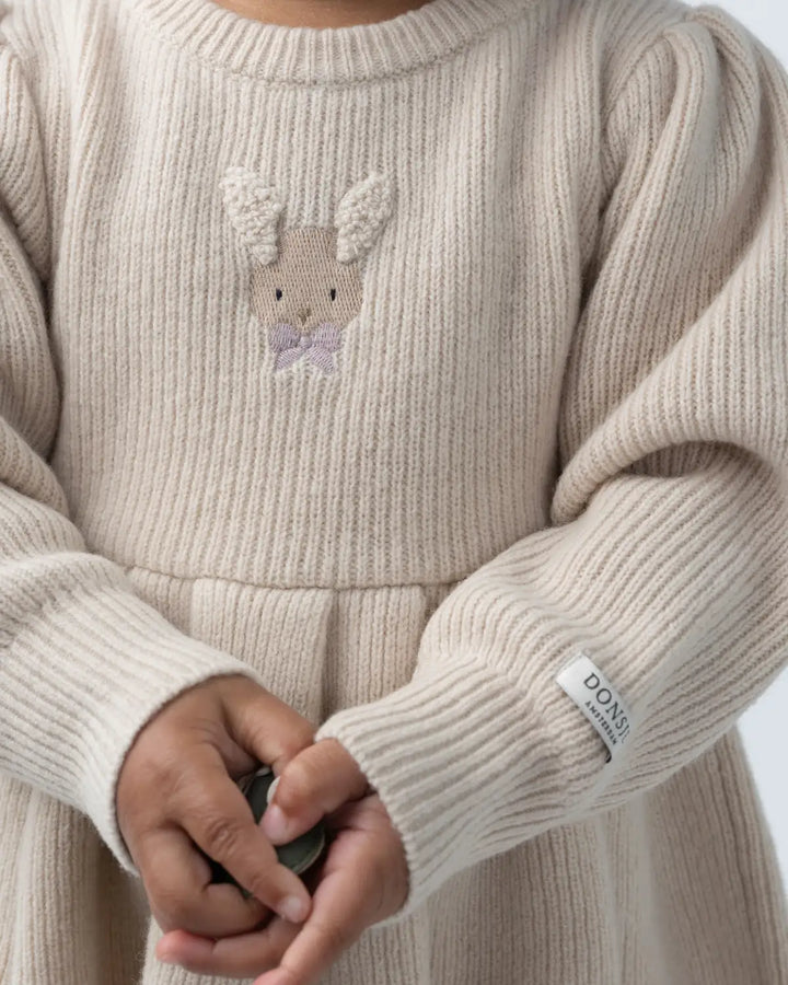 Donsje Vikkie Merino Wool Dess Fluffy Bunny Soft Sand getragen von einem Mädchen, Detailansicht aufgesticktes Hasengesicht und gerippte Strickstruktur