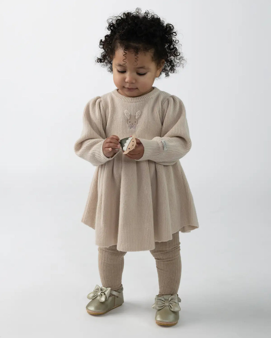 Donsje Vikkie Merino Wool Dess Fluffy Bunny Soft Sand getragen von einem Mädchen vor einer weißen Leinwand stehend, Ansicht von vorne. 