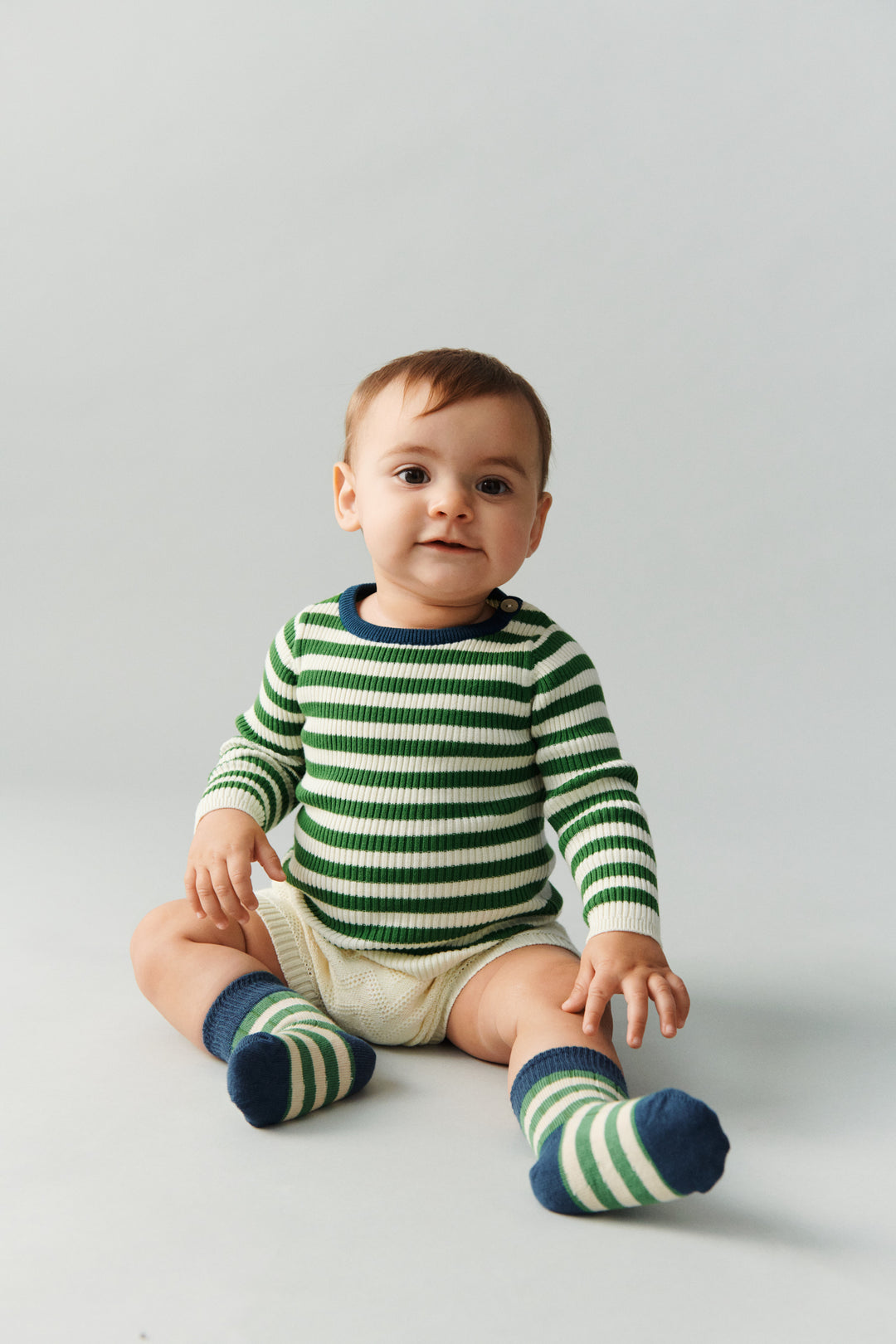 FUB Baby Rib Blouse ecru pine getragen von einem Baby sitzend vor weißem Hintergrund, kombiniert mit den Classic Strupe Socks in pine/ocean, Ansicht von vorne
