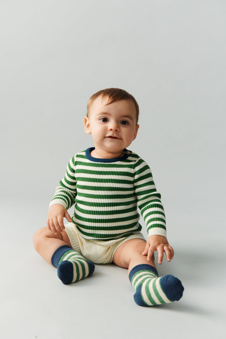 FUB Baby Rib Blouse ecru pine getragen von einem Baby sitzend vor weißem Hintergrund, kombiniert mit den Classic Strupe Socks in pine/ocean, Ansicht von vorne