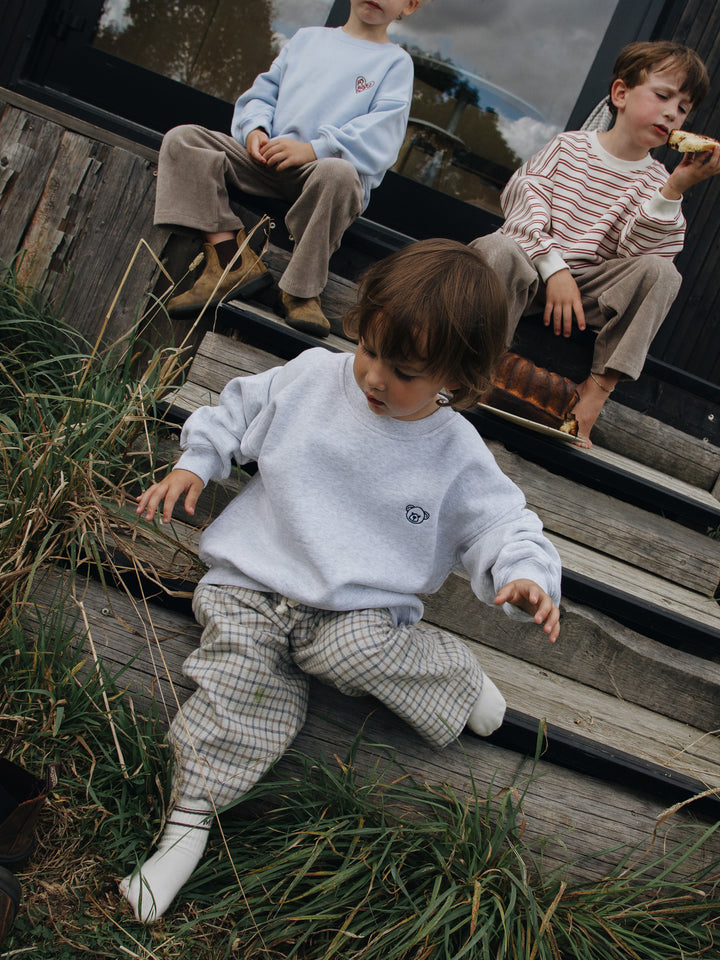 Famvibes Karo Pants Kids karo beige getragen von einem Jungen auf einer Treppe sitzend kombiniert mit dem Bear Sweatshirt Baby / Kids. 