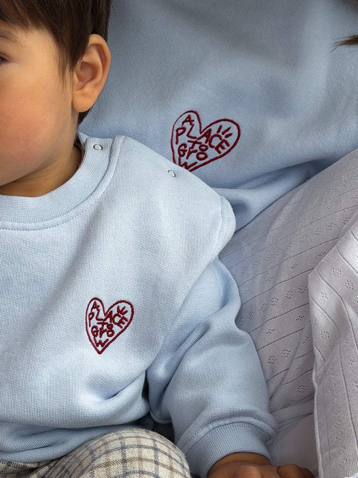 Famvibes Place To Grow Sweatshirt Baby/Kids blue getragen von einem kleinen Jungen vor seiner Mutter sitzend, die das gleiche Place To Grow Sweatshirt für Erwachsene trägt. 