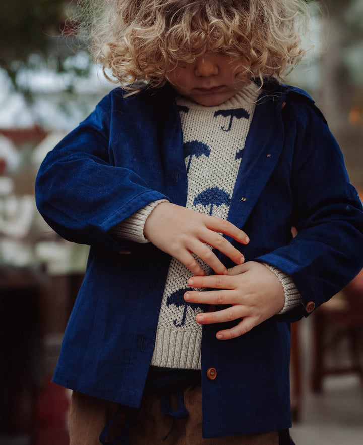 Fliink Janie Cashmere mbrella Pullover sandshell insignia blue getragen von einem Jungen in einem Gewächshaus stehend, Ansicht von vorne. Pullover befindet sich unter einem blauen Hemd. 