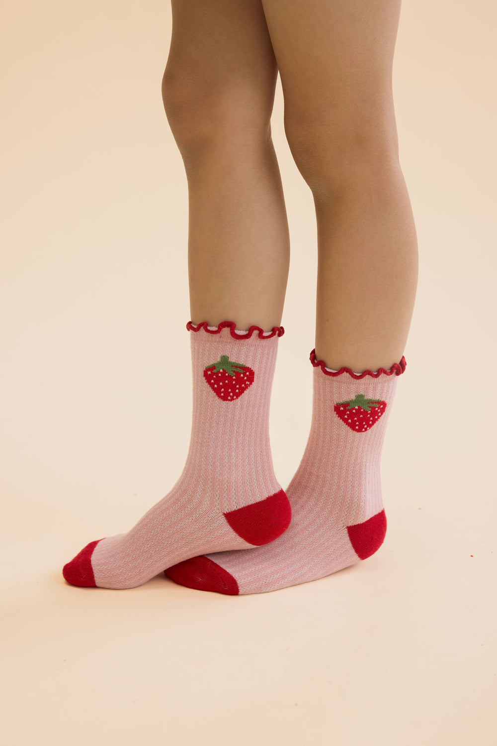 Floess_Vigga_Socks_2-pack_pointelle_berry_2-pack getragen von einem Mädchen vor hellgelbem Hintergrund, Fokus auf Socken, rosa Paar mit großen Erdbeeren 