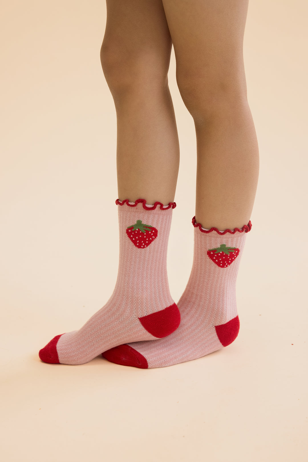 Floess_Vigga_Socks_2-pack_pointelle_berry_2-pack getragen von einem Mädchen vor hellgelbem Hintergrund, Fokus auf Socken, rosa Paar mit großen Erdbeeren 