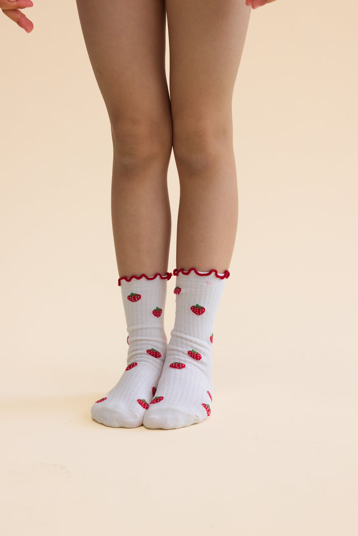 Floess_Vigga_Socks_2-pack_pointelle_berry_2-pack getragen von einem Mädchen vor hellgelbem Hintergrund, Fokus auf Socken. Weißes Paar mit kleinen Erdbeeren.