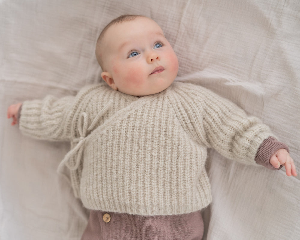 Halfen Alpaka Grobstrickjacke beige getragen von einem Baby auf einer Decke liegend, Ansicht von vorne oben. 