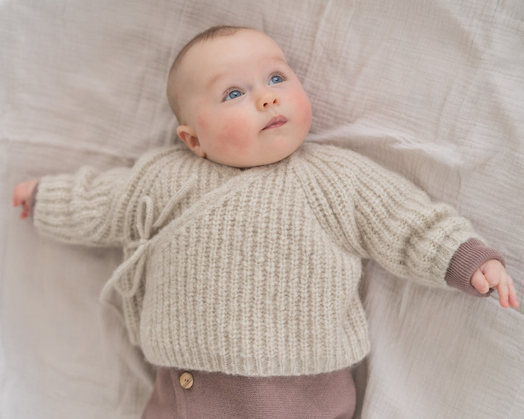 Halfen Alpaka Grobstrickjacke beige getragen von einem Baby auf einer Decke liegend, Ansicht von vorne oben. 