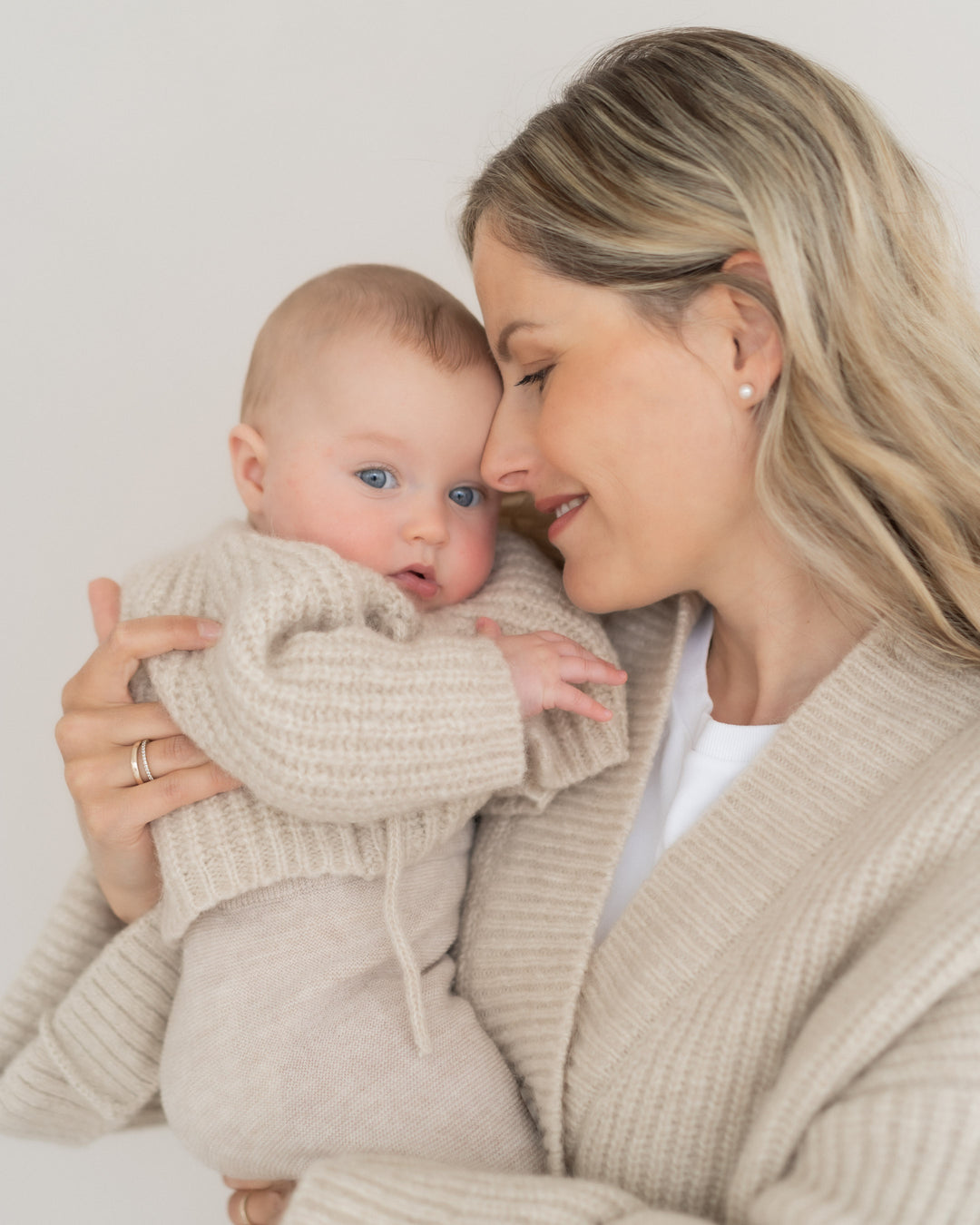 Halfen Alpaka Grobstrickjacke beige getragen von einem Baby gehalten von einer Frau, Ansicht von der Seite. 