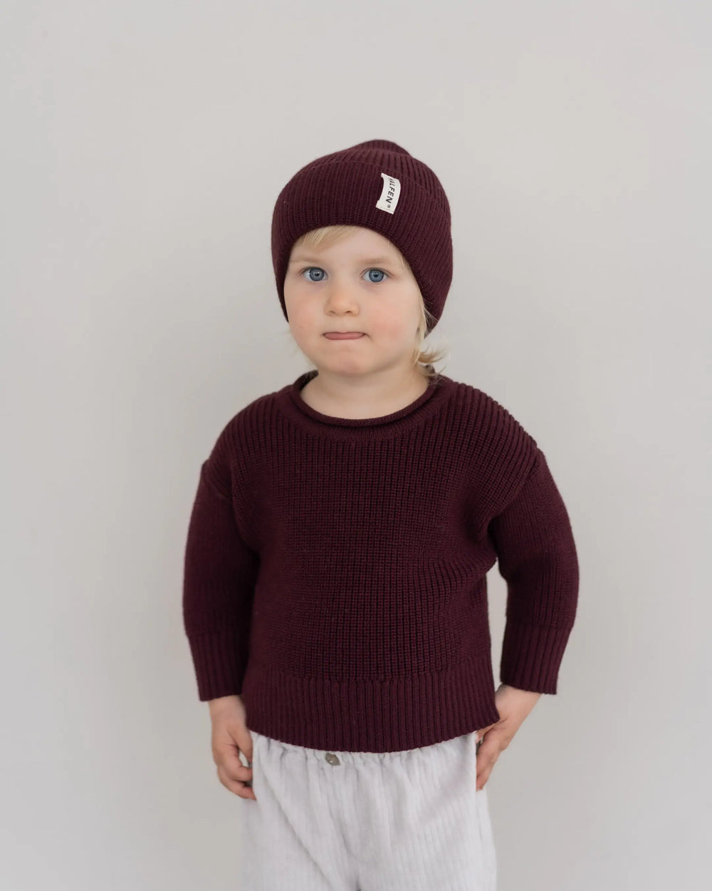 Halfen Strick-Beanie Merino cherry getragen von einem Kind vor beigefarbener Leinwand, Ansicht von vorne. 