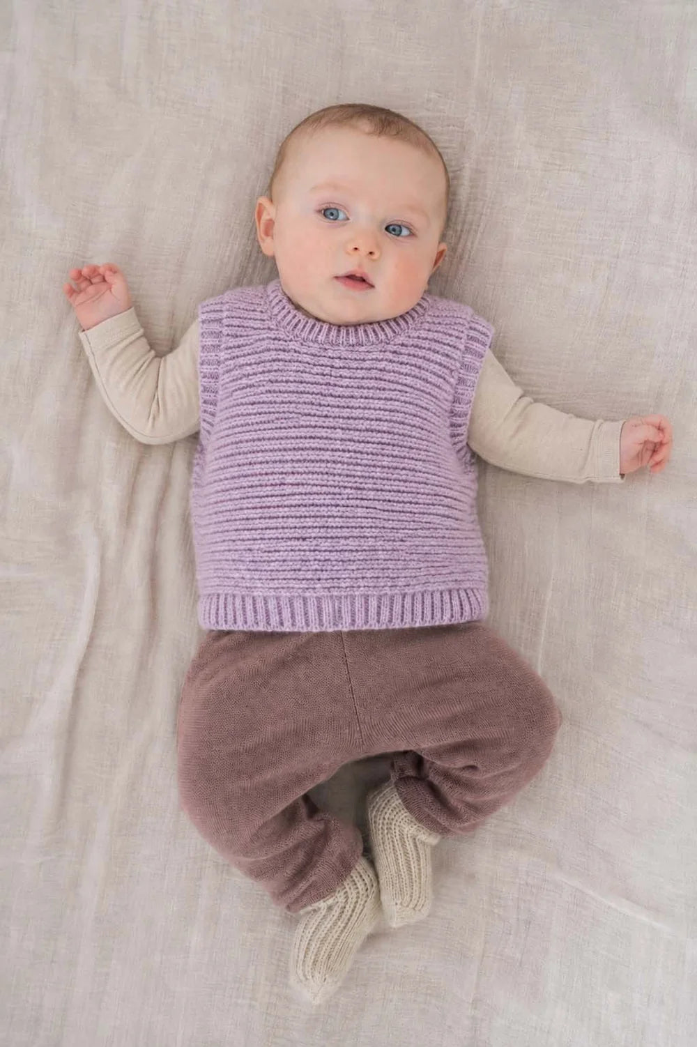 Halfen Struktur Pullunder Alpaka flieder getragen von einem Baby auf einem beigefarbenen Laken liegend, Ansicht von oben. 