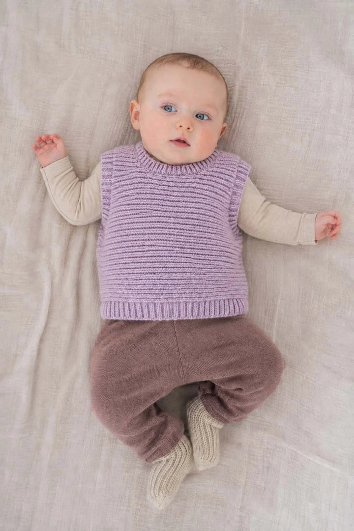 Halfen Struktur Pullunder Alpaka flieder getragen von einem Baby auf einem beigefarbenen Laken liegend, Ansicht von oben. 