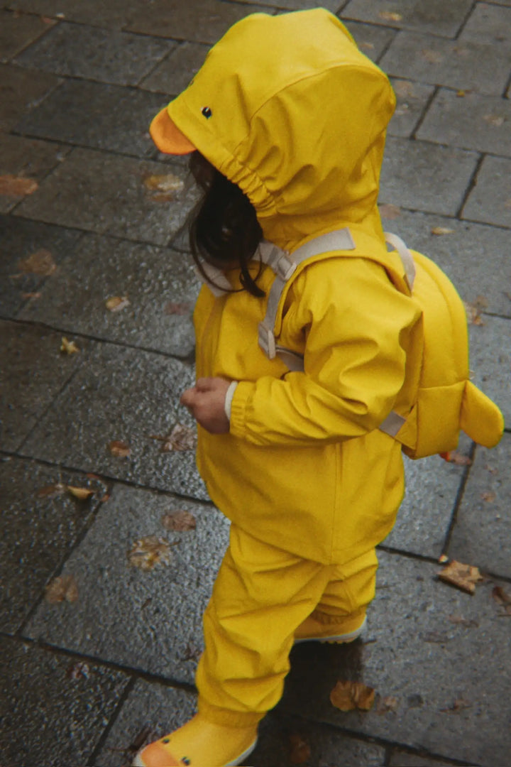 gelbe Regenjacke und -hose im Set für Kinder mit orangefarbenem Enten-Schnabel an der abnehmbaren Kapuze. Wasserdicht und windabweisend. Getragen von einem Mädchen, kombiniert mit den Anneli Regenstiefeln. 