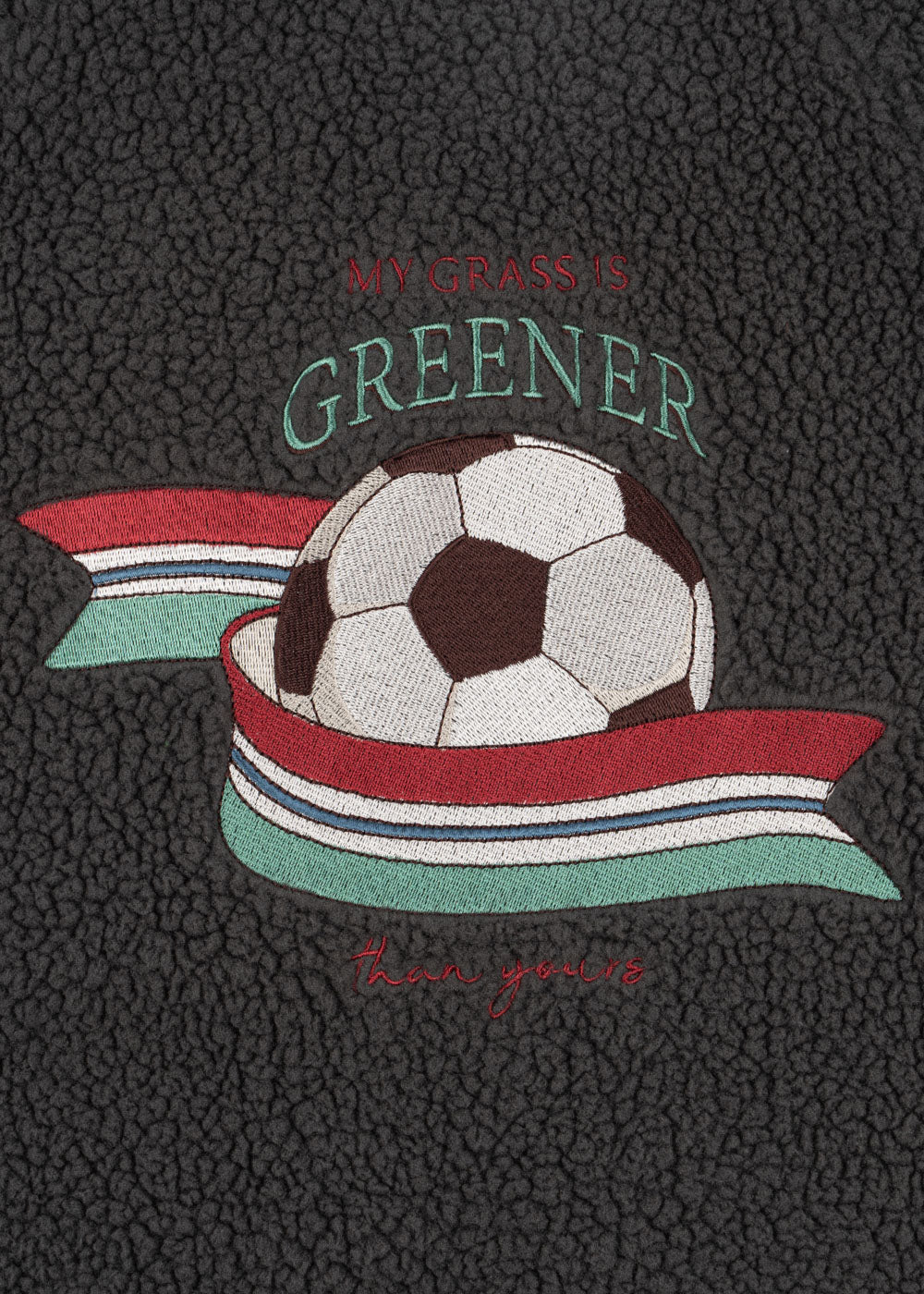 Konges Sloejd Jody Bomber Jacket GRS magnet Detailansicht aufgestickter Fußballd mit blau, weiß, rot und grüngestreiftem Band und dem Schriftzug "my grass is greener - than yours".