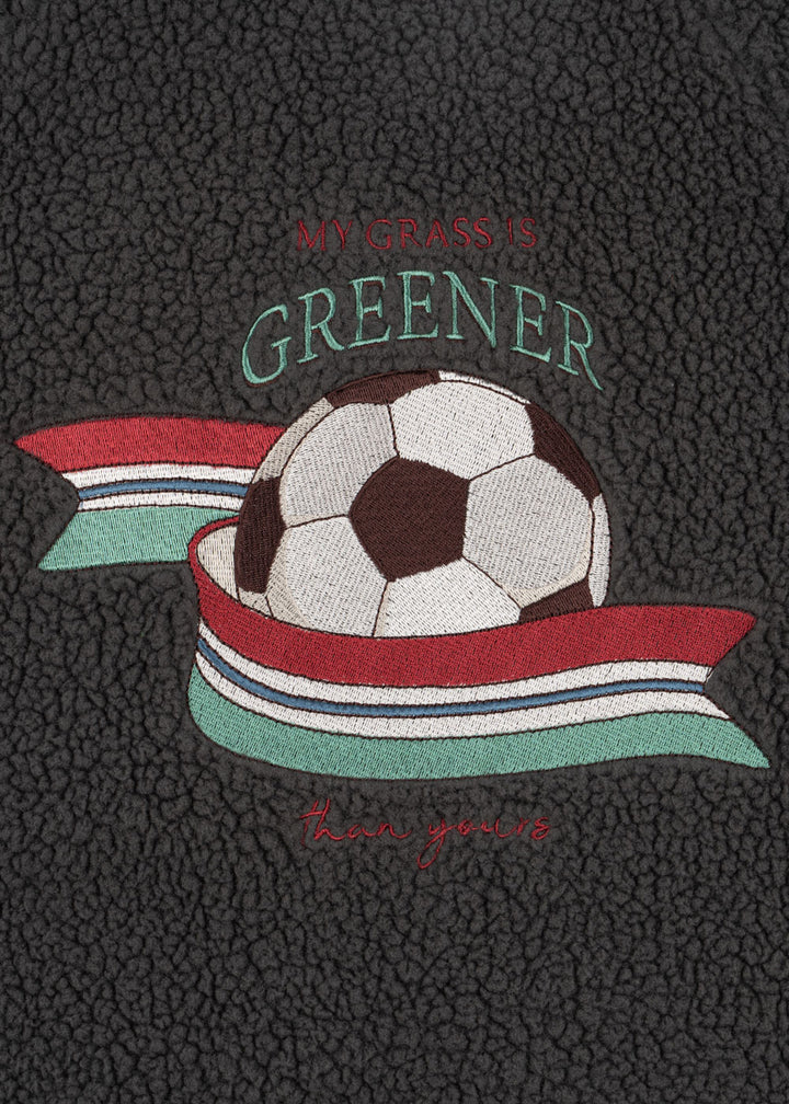 Konges Sloejd Jody Bomber Jacket GRS magnet Detailansicht aufgestickter Fußballd mit blau, weiß, rot und grüngestreiftem Band und dem Schriftzug "my grass is greener - than yours".