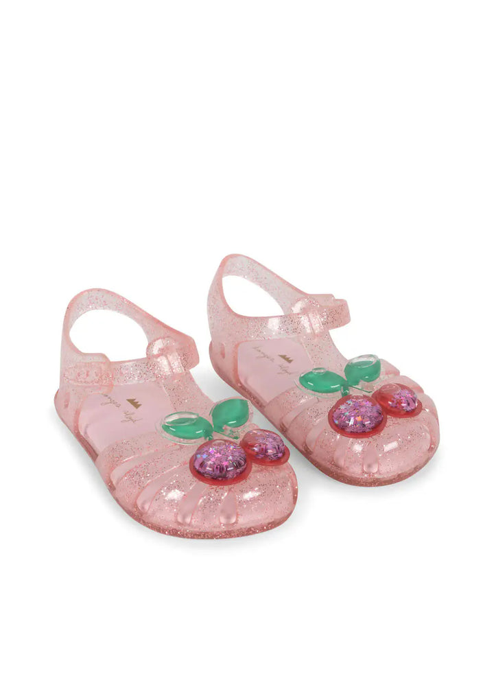 rosafarbene Sandalen für Mädchen aus glitzerndem PVC mit Riemchen-Klettverschluss und einer großen mit Glitzer-Gel gefüllten Kirsche vorne auf dem Fuß. Produktbild, Ansicht von vorne