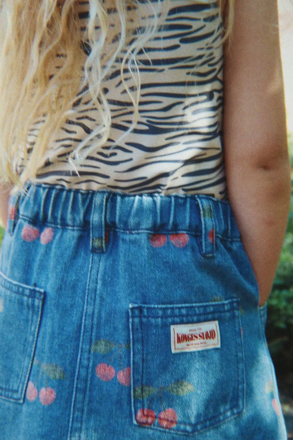 Konges Sloejd Magot Printed Skirt GOTS ma grande cerise denim getragen von einem Mädchen, Ansicht der Rückseite, zwei Gesäßtaschen, Kirschprint und Logo-Aufnäher