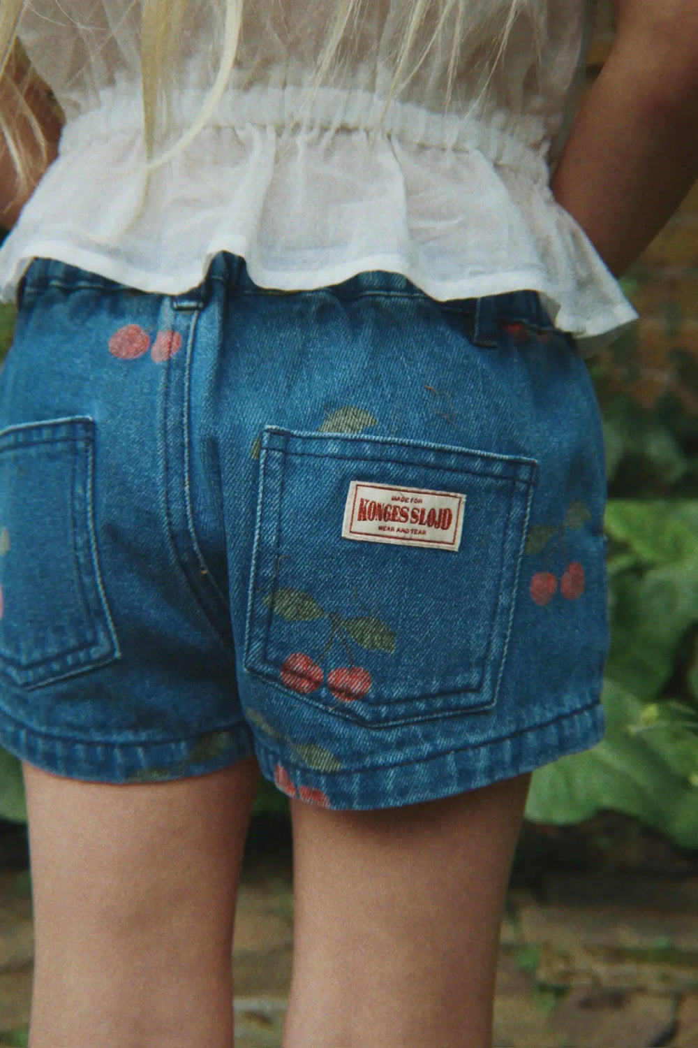 Konges Sloejd Magot Shorts GOTS ma grande cerise denim getragen von einem Mädchen, Ansicht der Rückseite, zwei Gesäßtaschen, Kirschprint und Konges Sloejd Logo