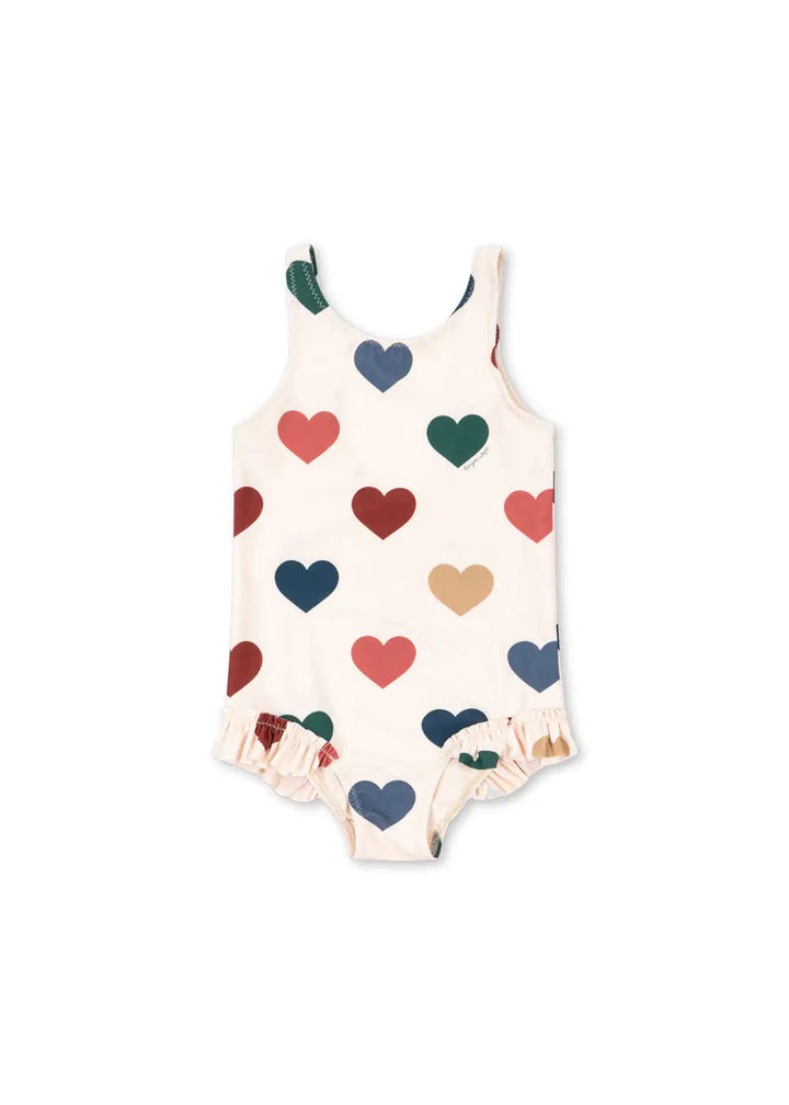cremefarbener Badeanzug für Mädchen mit buntem Herz-Allover-Print und Rüschen an den Beinausschnitten. Produktbild, Ansicht von vorne.