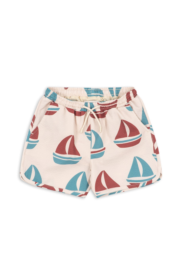cremefarbene Badeshorts für Jungen aus Polyester mit blau-rotem Segelboot-Allover-Print, Gummibund, verstellbarer Kordel, seitlichen Eingriffstaschen. Produktbild, Ansicht von vorne