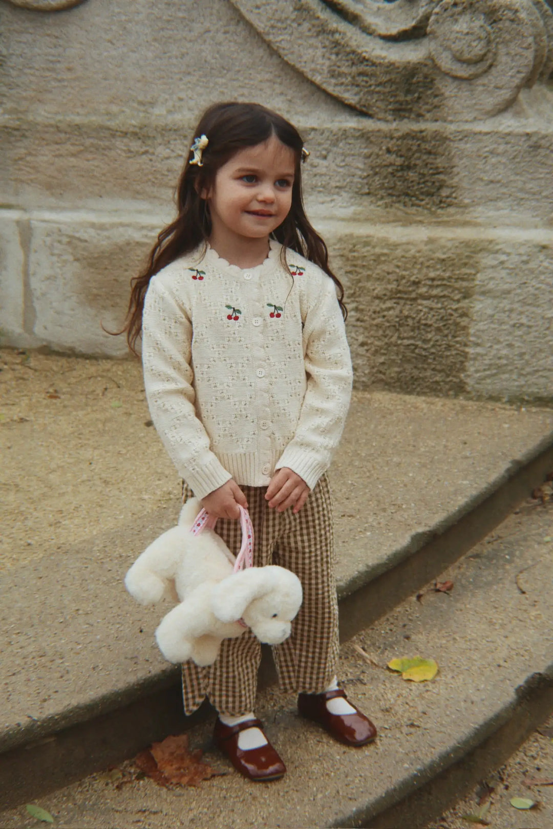 Konges Slojd Cocolo Cardigan buttercream getragen von einem kleinen Mädchen auf einer Brücke stehend, Ansicht von vorne. 