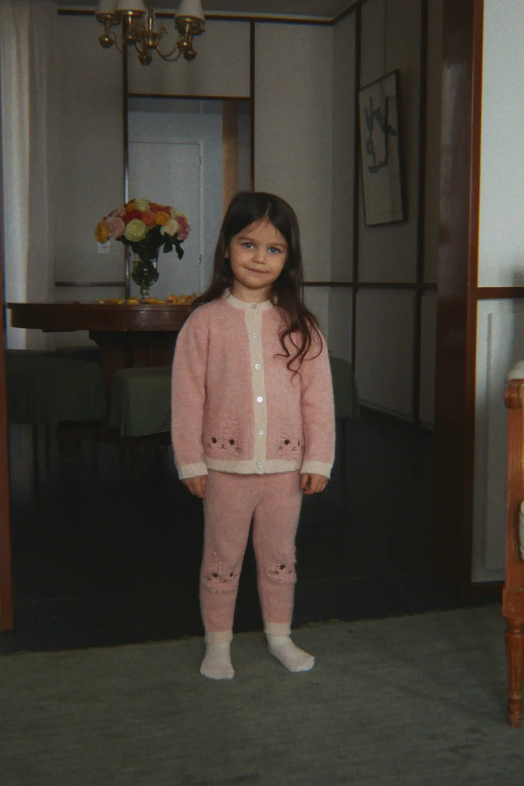 Konges Slojd Kitty Cardigan silver pink getragen von einem kleinen Mädchen, das in einem Türrahmen steht, Ansicht von vorne. 