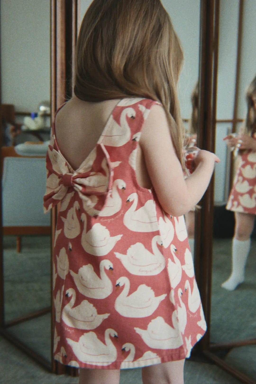 Konges Slojd Magot Bow Dress GOTS swan rose getragen von einem kleinen Mädchen vor einem Spiegel stehend, Ansicht der Rückseite. 
