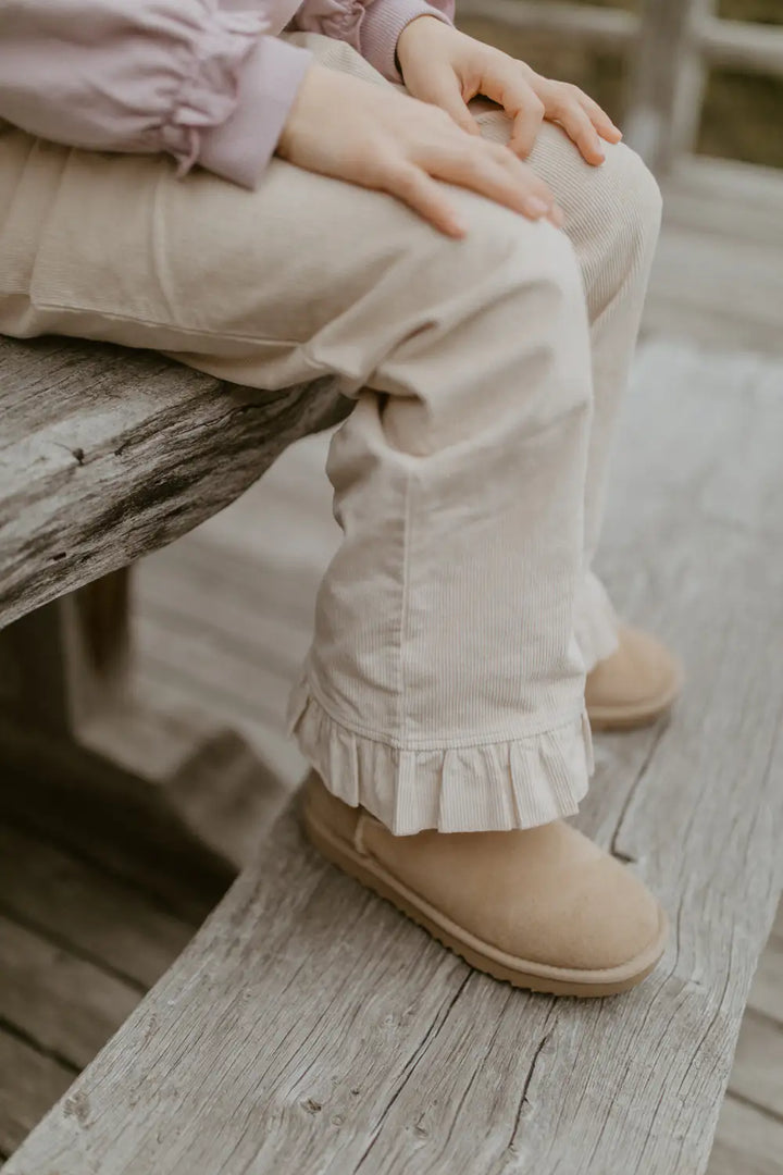 beigefarbene Cordhose aus Baumwolle und Elasthan für Mädchen mit geradem Bein, Rüschen am Saum der Fußenden und mit breitem bequemem Gummibund. Getragen von einem Kind, Detailansicht Rüschen am Fußende. 