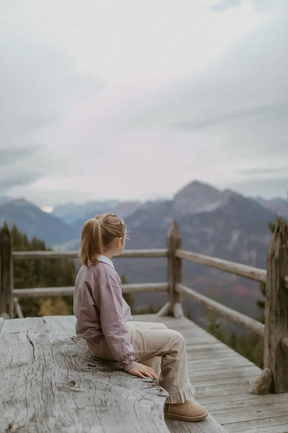 Leevje Cordhose mit Rüschen dusty rose getragen von einem Mädchen vor einer Berglandschaft sitzend, Ansicht von der Seite.