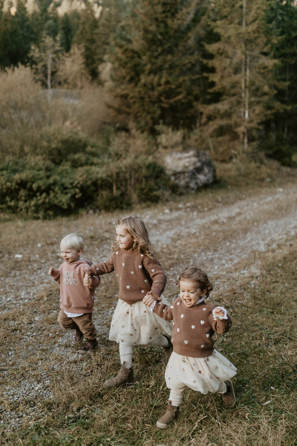 Leevje Mini-Me Chunky-Strickpullover Hearts Kinder pecan getragen von zwei Mädchen im Wald laufend, Ansicht von vorne. 