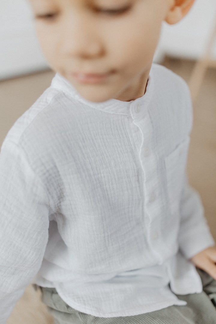 weißes Musselin-Hemd für Jungen aus 100% Baumwolle mit Stehkragen, Brusttasche auf der linken Seite und durchgehender Knopfleiste, getragen von einem Jungen