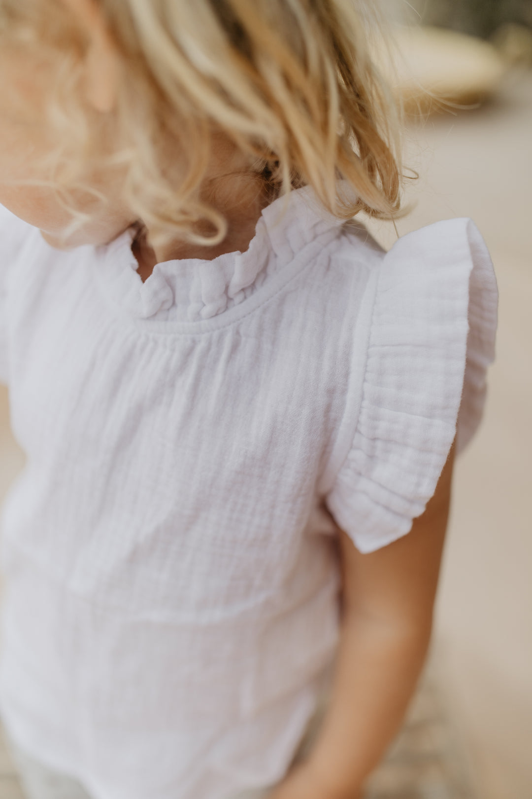 weißes Musselin-Top für Mädchen aus 100% Bio-Baumwolle mit gerüschtem Stehkragen und Rüschen an den Armausschnitten, getragen von einem Mädchen, Fokus auf Rüschen am Armausschnitt