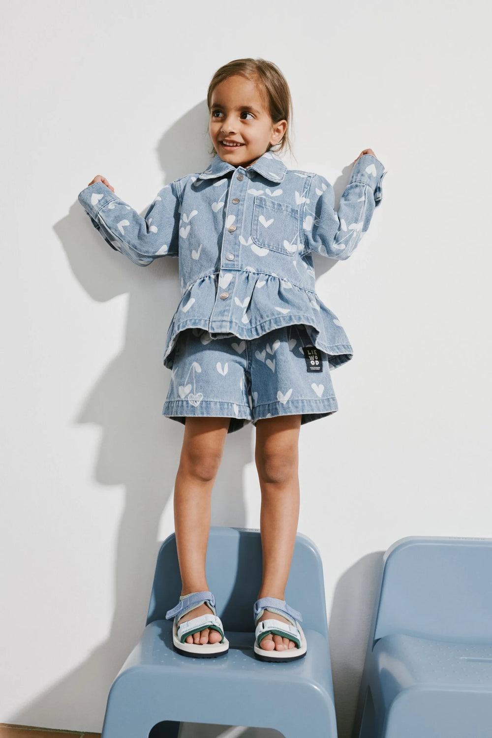 Liewood_Kenza_Printed_Denim_Overshirt sweethearts light blue denim getragen von einem Mädchen, das auf einem Stuhl vor einer weißen Wand steht und die stilgleiche Shorts zu dem Overshirt trägt