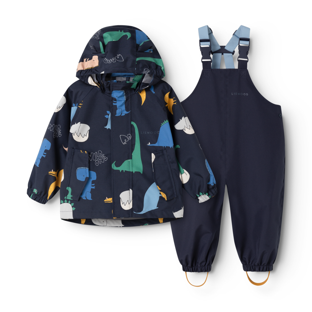 Set aus Regenjacke und Regenhose für Kleinkinder. Dunkelblaue Regenjacke mit buntem Dino-Allover-Muster, Kapuze, kleinen Ohren an der Kapuze, durchgehender Reißverschluss. Dunkelblaue Regenhose mit verstellbaren Hosenträgern sowie Schlaufen an den Fußenden. Reflektierendes Emblem am Rücken der Jacke und am hinteren Hosenbein.