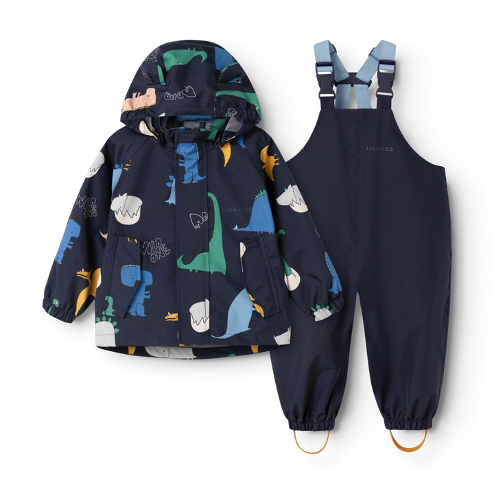 Set aus Regenjacke und Regenhose für Kleinkinder. Dunkelblaue Regenjacke mit buntem Dino-Allover-Muster, Kapuze, kleinen Ohren an der Kapuze, durchgehender Reißverschluss. Dunkelblaue Regenhose mit verstellbaren Hosenträgern sowie Schlaufen an den Fußenden. Reflektierendes Emblem am Rücken der Jacke und am hinteren Hosenbein.