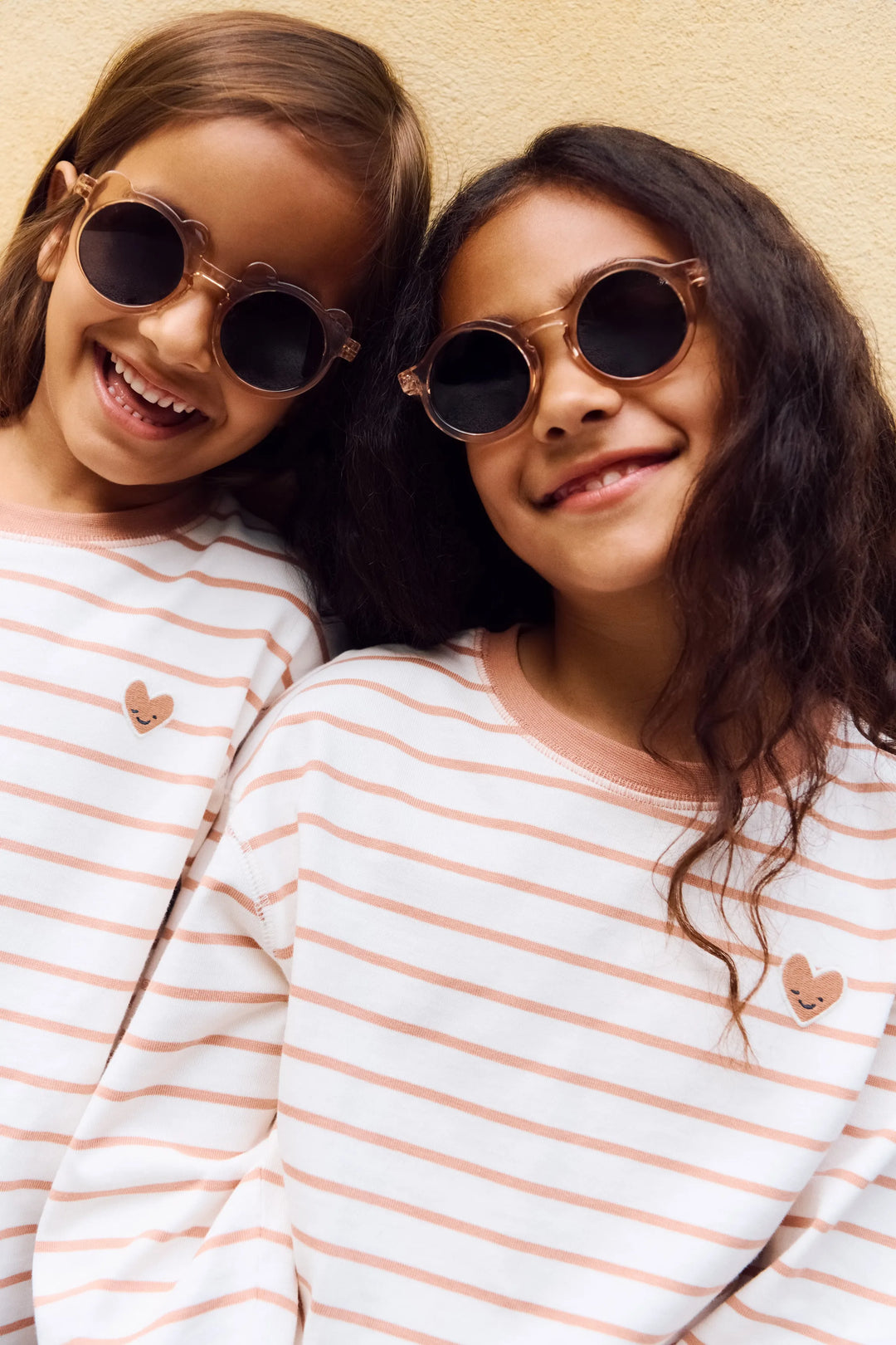 Liewood Tyler Stripe LS t-shirt heart tuscany rose / creme de la creme getragen von zwei nebeneinander stehenden Mädchen vor einer gelben Mauer mit Fokus auf die Langarmshirts